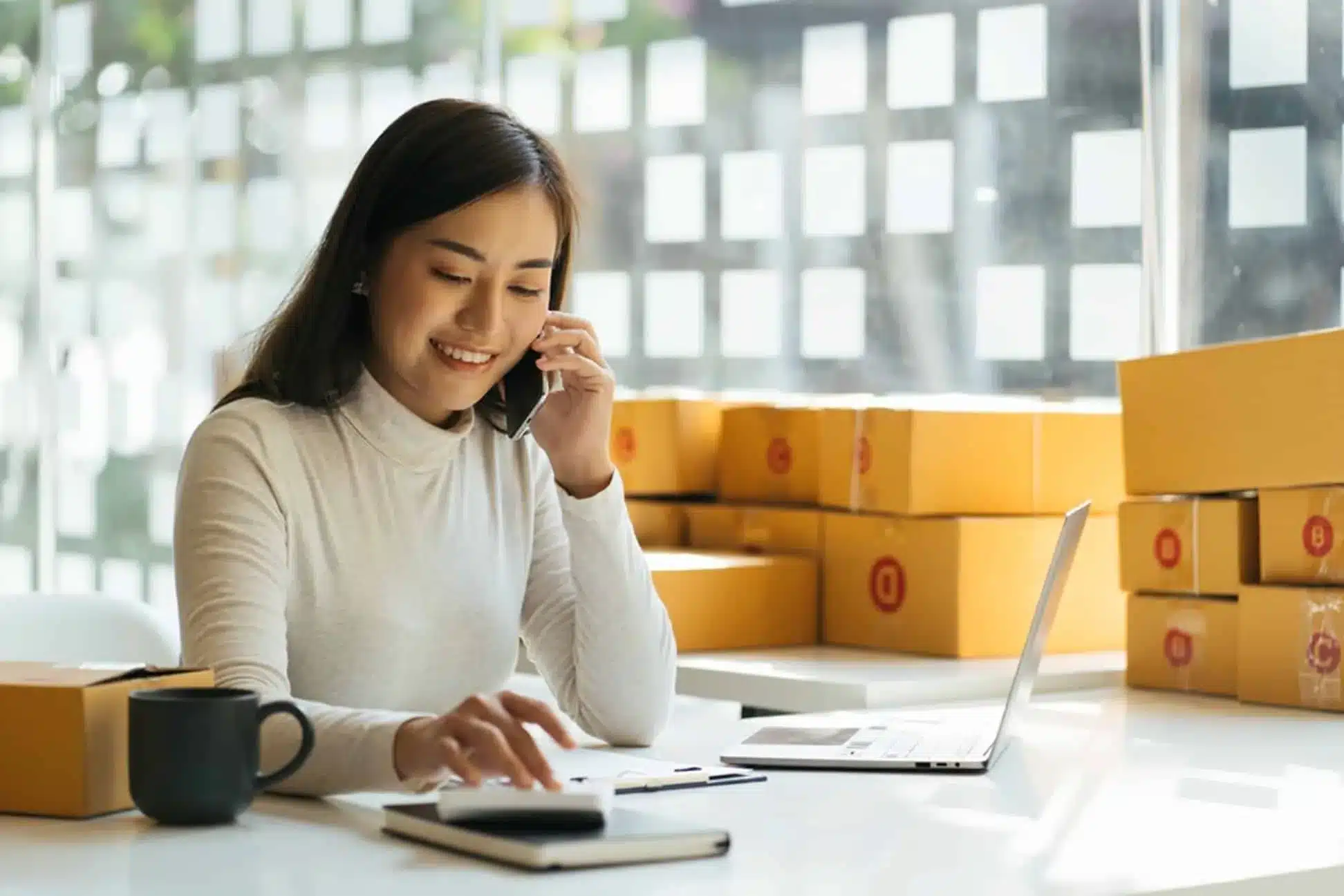 Woman managing supply chain operations on laptop with shipping boxes nearby