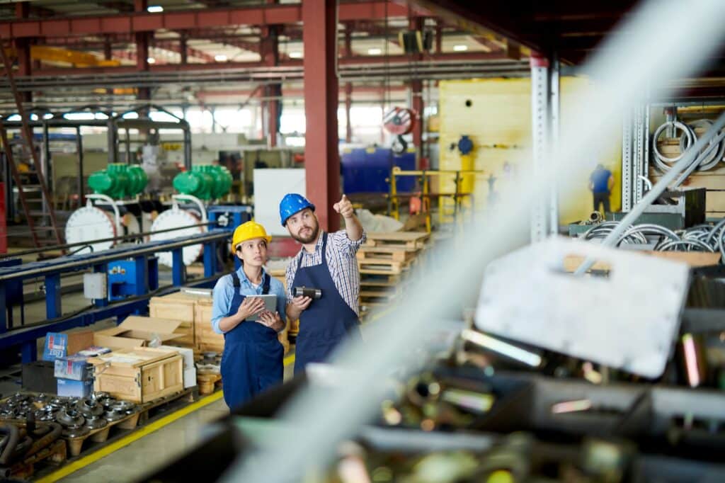 Two factory workers in hard hats reviewing production data on tablet in manufacturing facility