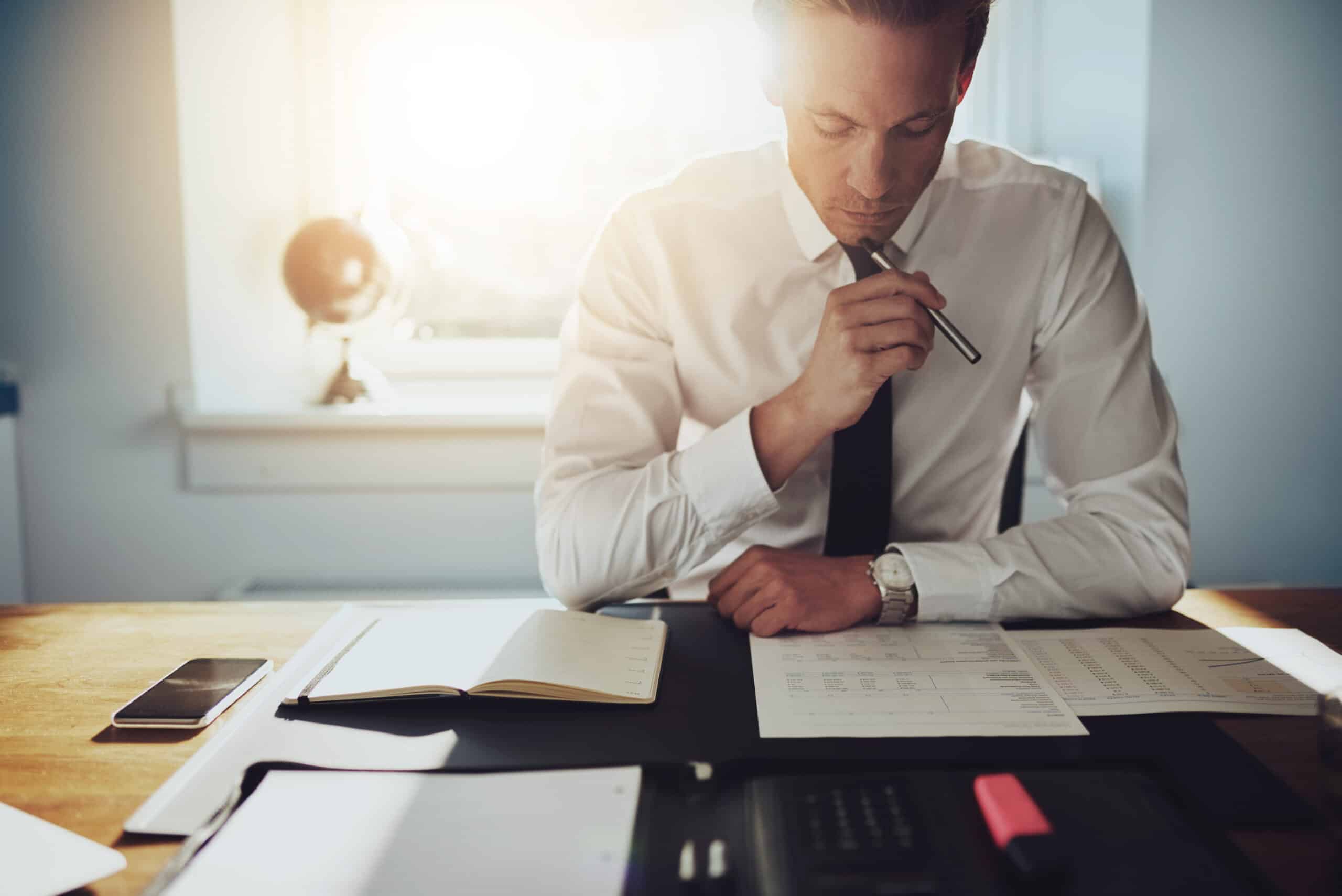 Businessman reviewing a spreadsheet report at his desk, holding a pen in thought