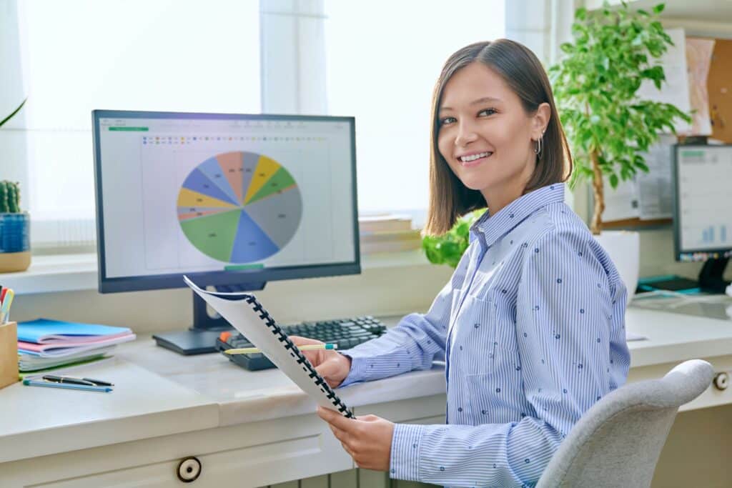 a woman smiling at the camera with a laptop showing charts on her desk behind her